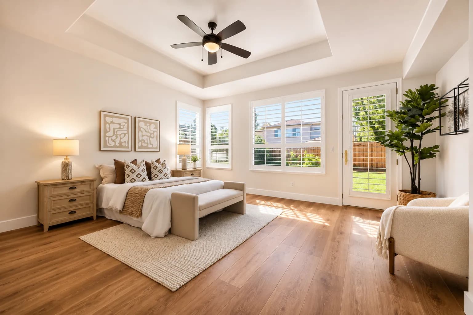 Staged primary bedroom with a tray ceiling, light wood dresser and doors to the garden