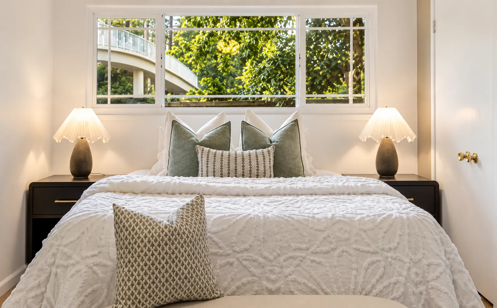 Staged bedroom with white textured bedding, sage pillows and scalloped lamps under a garden window