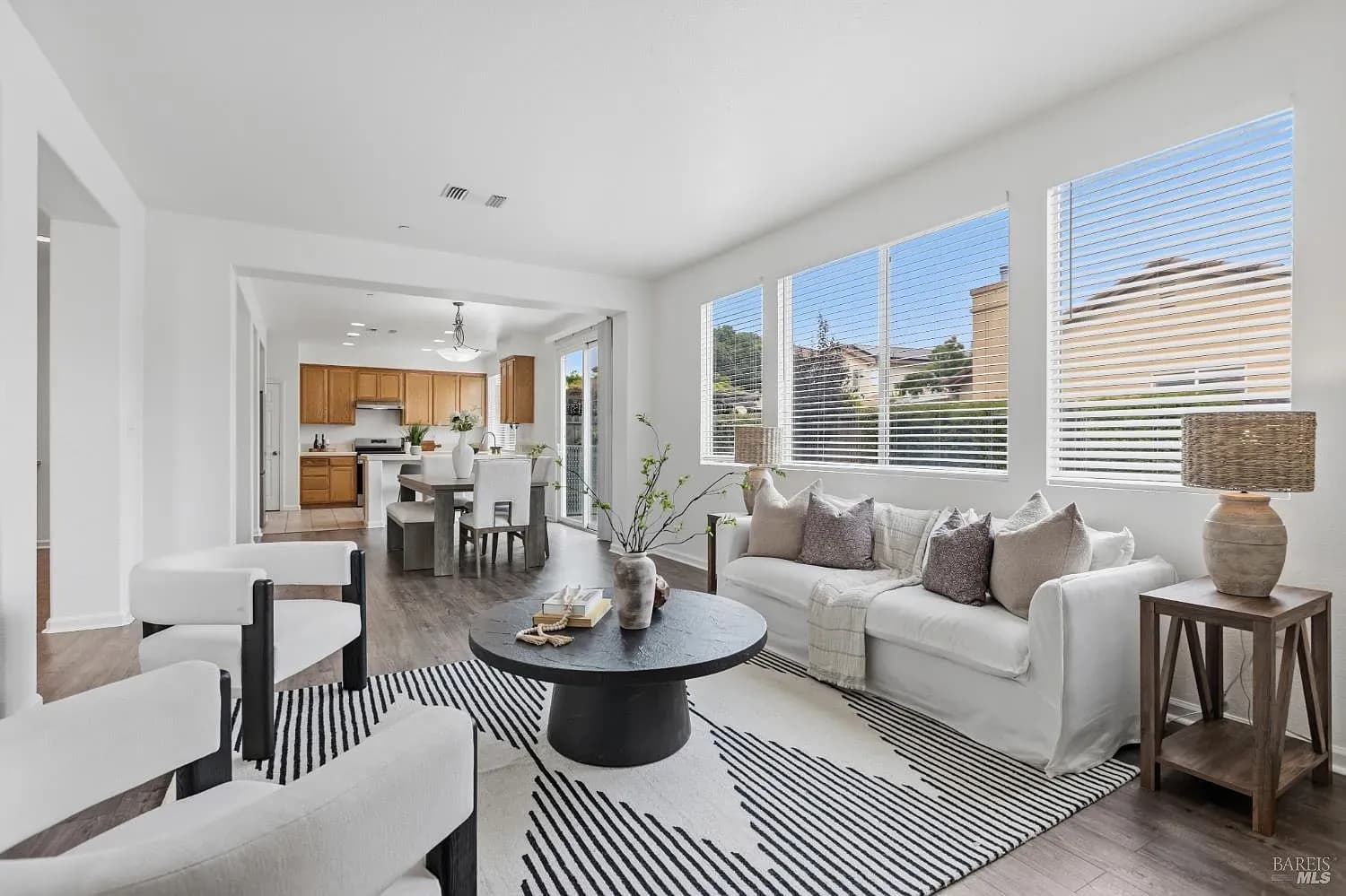 Staged living room with a white sofa, a black round coffee table and a striped rug, kitchen beyond