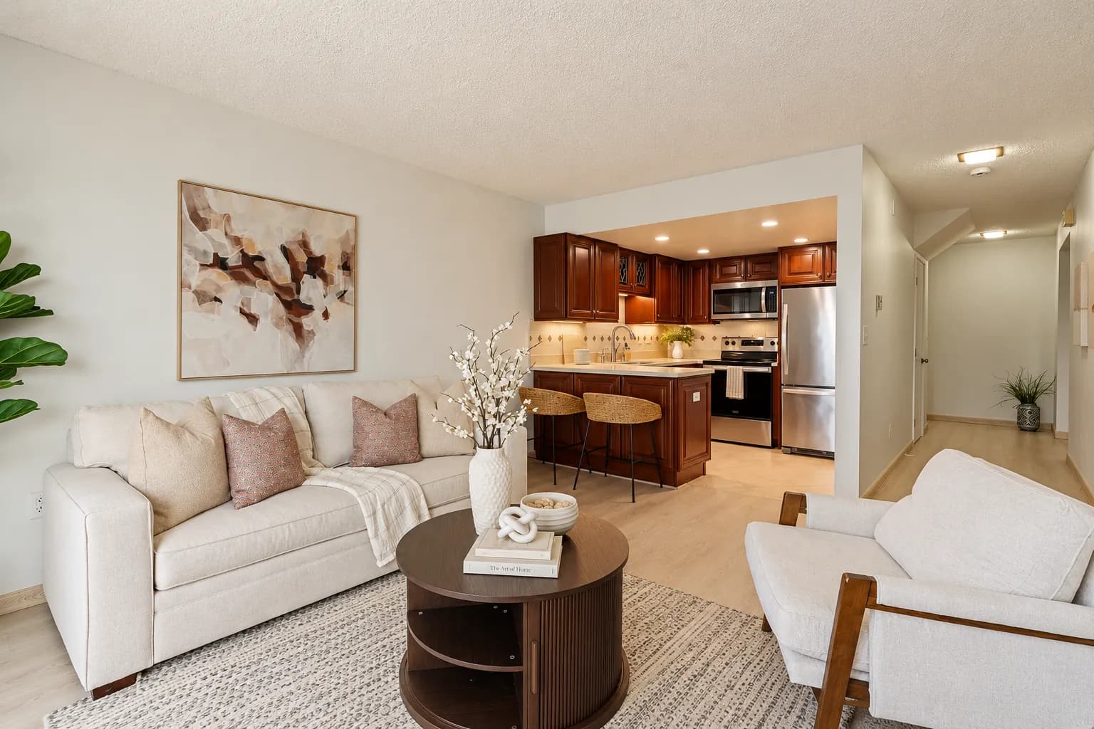 Staged Daly City living room with a white sofa, blush pillows and a round wood coffee table, kitchen beyond